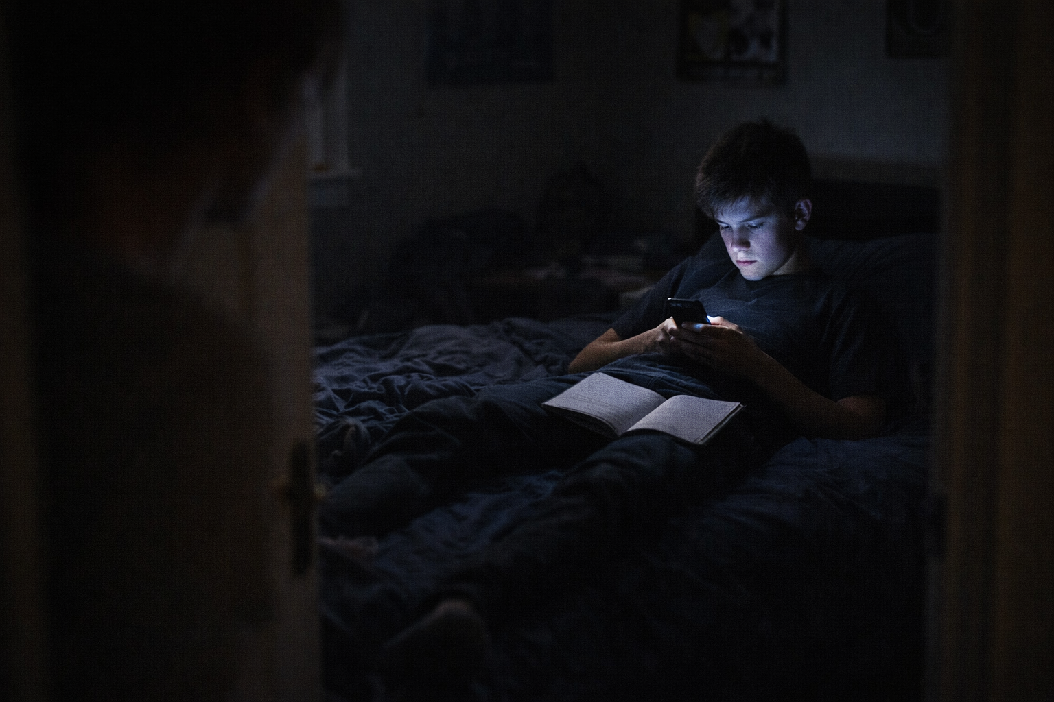 Modra svetloba zaslona osvetljuje tiho razdaljo med staršem in najstnikom, medtem ko šolski zvezek ostaja odprt, a prezrt.